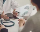 Lekarz w białym kitlu rozmawia z pacjentką w gabinecie. Na stole stetoskop i laptop. S1 Norwegia.