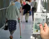 Emerytka z laską idzie korytarzem, obok stos banknotów 100 zł. Waloryzacja emerytur 2026 - tabela.