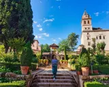 Frau spaziert durch einen üppigen Garten mit Zypressen und einem Turm im Hintergrund. Ein schöner Ort für einen Tagesausflug, vielleicht sogar von Malaga aus.