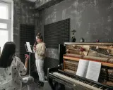 Dziewczynka ćwiczy swój **alt głos** przy mikrofonie, podczas gdy nauczycielka słucha jej uważnie. Obok stoi pianino z nutami.