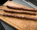 Drei saftige Adana Kebap Spieße liegen auf einem Holzbrett. Perfekt für ein authentisches Adana Kebap Rezept.