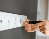 Ręka z wkrętakiem przy białym gniazdku. Dowiedz się, jak przenieść gniazdko bez kucia, wykorzystując nowoczesne rozwiązania.
