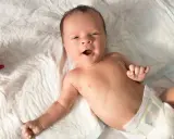 Baby mit offenem Mund, vielleicht im 6-Wochen-Schub, liegt auf einer weißen Unterlage neben einem bunten Spielzeug.