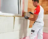Pracownik montuje wełnę mineralną, wykonując ocieplenie budynku. Widać okno i materiały izolacyjne.