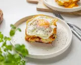 Pyszny croque madame z jajkiem sadzonym na wierzchu, na talerzu, obok chleba i zielonych liści.
