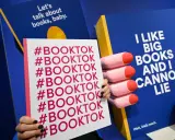 Mani che tengono un cartello con #BOOKTOK ripetuto. Sullo sfondo, un libro blu con la scritta "I LIKE BIG BOOKS AND I CANNOT LIE".
