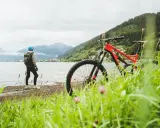 En cyklist med hjälm och ryggsäck står vid en sjö med berg i bakgrunden. En mountainbike står redo för äventyr på mtb leder.