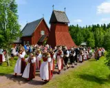 En procession av musiker i folkdräkter, som spelar fiol och gitarr, går förbi en röd träkyrka. Många människor i traditionella kläder, vilka firar midsommar, följer efter.