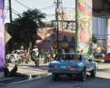 Kolorowe murale zdobią filary wiaduktu w słoneczny dzień. Na ulicy ruchliwie: ludzie na motocyklach i quadach, samochody, psy. To scena z GTA 6 online.
