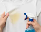 Manos aplicando spray a una mancha amarilla en camiseta blanca. Aprende como quitar boli de la ropa con este método.
