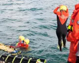 Sauvetage en mer : une sauveteuse en combinaison noire et orange se prépare à plonger. Deux personnes en gilets de sauvetage et casques jaunes flottent près d'une civière. Communication VHF et MMSI essentielle.