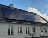 Ett hus med svarta, glänsande nya solceller på taket. Solen reflekteras i panelerna mot en blå himmel med moln.