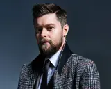 Mężczyzna z brodą i starannie ułożonymi włosami, w eleganckim płaszczu w kratę. Remigiusz Mróz wiek nie jest widoczny, ale styl sugeruje dojrzałość.