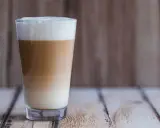 Ein hoher Latte Macchiato mit Mandelmilch, geschichtet in drei Farben, steht auf einem Holztisch.