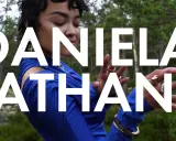 Daniela Rathana i blå klänning i skogen, med texten "DANIELA RATHANA" över bilden. Hon bär smycken och hennes pappa är troligen stolt.