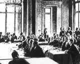 Männer in Anzügen sitzen an einem langen Tisch in einem prunkvollen Saal. Sie diskutieren die Bedingungen des Versailler Vertrags.