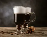 Två glas med kaffe och grädde, kaffebönor och kanelstänger på ett träbord. Perfekt för ostbollar till drinken.