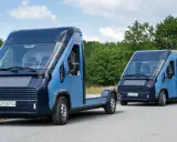 Dwa nowoczesne, niebieskie polskie samochody elektryczne na drodze wśród zieleni. Jeden to podwozie, drugi to furgonetka.