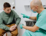 Lekarz w zielonym fartuchu i masce rozmawia z młodym pacjentem, wskazując palcem. Młody mężczyzna ma zmartwioną minę, być może z powodu obrzęku penisa.