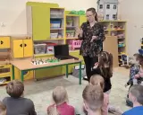Nauczycielka prowadzi zajęcia kreatywne dla dzieci. Na stole przed nimi leżą klocki Lego, a dzieci z uwagą słuchają.