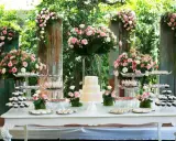 Mesa de dulces con pastel de tres pisos, cupcakes y postres variados, decorada con abundantes flores rosas y blancas. Ideal para hacer flores de golosinas.