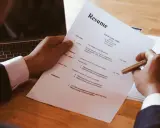 Dłoń trzymająca długopis nad dokumentem CV obok laptopa.