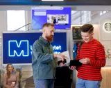 Dwóch studentów dyskutuje o kierunkach studiów WSB, trzymając laptopa i tablet. W tle widoczna jest neonowa litera "M" i ekran informacyjny.