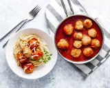 Pyszne pulpety w sosie pomidorowym z makaronem spaghetti, posypane parmezanem. Obok patelnia z gorącymi pulpetami w sosie.