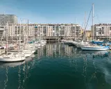 Barche a vela ormeggiate nel porto di Tolone, con edifici residenziali sullo sfondo e il cielo azzurro.