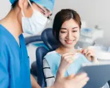 Uśmiechnięta pacjentka trzyma przezroczyste nakładki, a dentysta w maseczce i czepku pokazuje jej coś na tablecie. To pokazuje, jak zwracać się do dentysty.