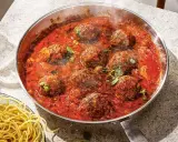 Frikadeller i tomatsås som puttrar i en panna, med en sidokorg med spaghetti.