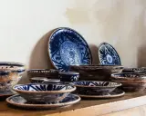 Ceramiche artigianato toscano con decori blu su fondo grezzo. Piatti e ciotole disposti su una mensola di legno.