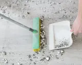 Main d'une personne utilisant un balai et une pelle pour essuyer les plâtres et débris sur un sol en bois.