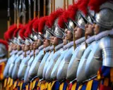 Guardie svizzere in fila, con elmi e piume rosse. Curiosità sulla loro uniforme storica e il loro ruolo.