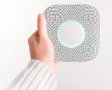 En hand håller upp en vit Google Nest Protect-rökdetektor med blåaktig glöd.