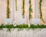 Stół weselny w stylu greenery, udekorowany bujną zielenią, białymi różami i świecami. Tło tworzą zwiewne zasłony i delikatne światełka.