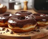 Un beignet au chocolat, généreusement glacé, repose sur une planche en bois. Des pépites de chocolat parsèment la scène.