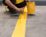 Persona pintando una línea amarilla sobre suelo de cemento sin pulir, usando una brocha y un cubo de pintura.