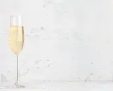 Ett glas champagne med bubblor, kanske en Jaques Selosse, står på ett vitt bord mot en ljus, texturerad bakgrund.