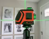 Pomarańczowa poziomica laserowa z zielonymi liniami na statywie, na tle oprawionych zdjęć.