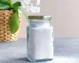 En glasburk med vitt, kornigt eget maskindiskmedel står på en grå yta. En korg med en orkidé och vita blommor syns till vänster.