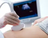 Ręka w rękawiczce trzyma sondę USG, która bada brzuch. Na ekranie widać obraz z badania, być może to test pappa.