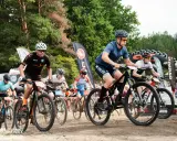 Kolarze biorą udział w wyścigu **zawody MTB**. Widać ich w akcji, jadących po piaszczystej trasie, z lasem w tle.