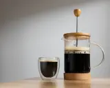Frisch gebrühter Kaffee mit Wasser in einer French Press und einem Glas. Der Duft von Kaffee liegt in der Luft.