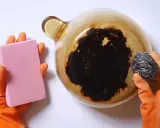 Manos con guantes naranjas sostienen una esponja rosa y un estropajo metálico, listos para aprender como quitar el quemado de una olla.