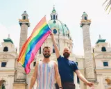 Two men proudly hold a rainbow flag in front of a grand building in Austria, celebrating gay pride.