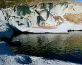 Scogliere bianche erose dal mare, con vegetazione secca e un'acqua scura che riflette il cielo. Sembra un luogo selvaggio, quasi un "cane malu" che custodisce la costa.