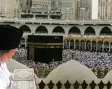 Chłopiec w czapce i białej szacie czyta Koran na tle tłumu pielgrzymów i Kaaby w Mekce.