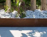 Parterre devant maison sans entretien, avec des galets blancs, des piquets en bois et une bordure rouillée. Des fleurs blanches éclosent.