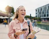 Szczęśliwa studentka z polską maturą, która planuje studia za granicą, śmieje się, trzymając telefon i książki.